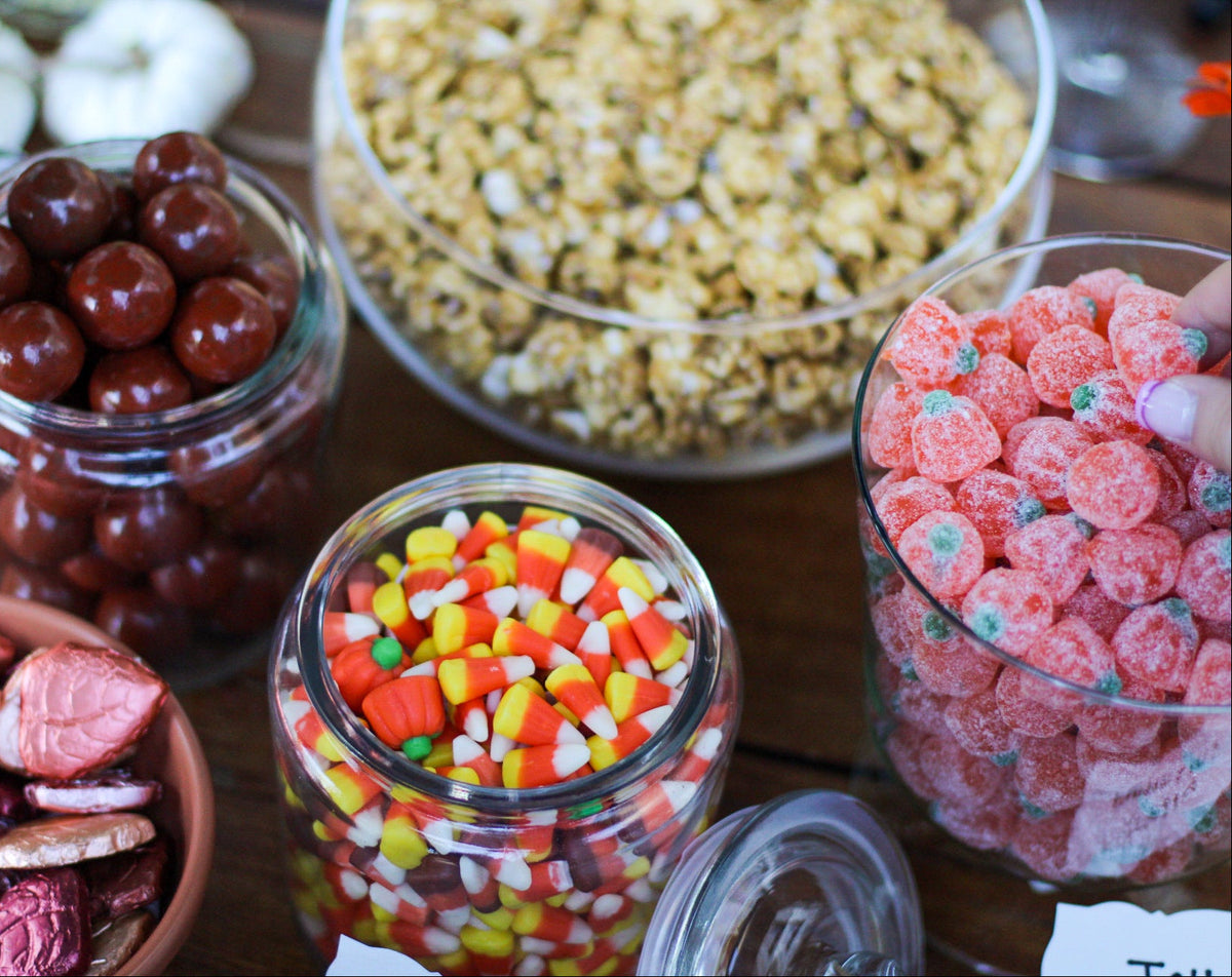 A close-up of a candy display with several glass jars and bowls on a wooden table. Visible items include dark red round candies, candy corn, pink strawberry-shaped candies, heart-shaped chocolates, and a bowl of caramel popcorn.