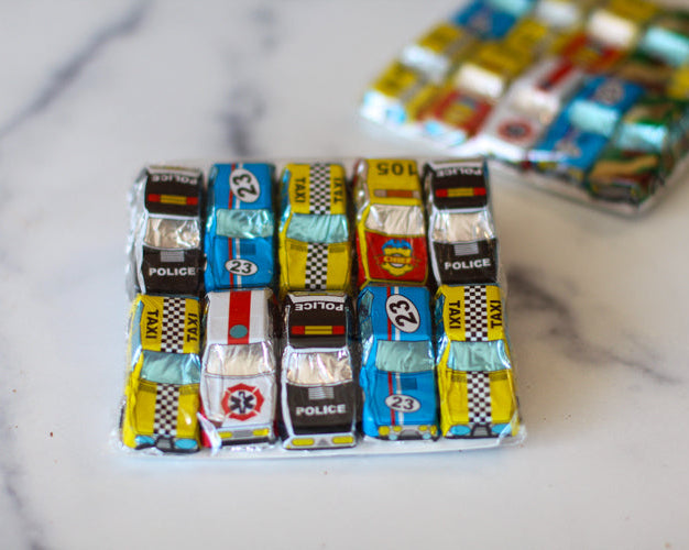 Mini chocolate bars shaped like cars, wrapped in colorful foils with racing and emergency vehicle patterns, displayed on a marble surface.