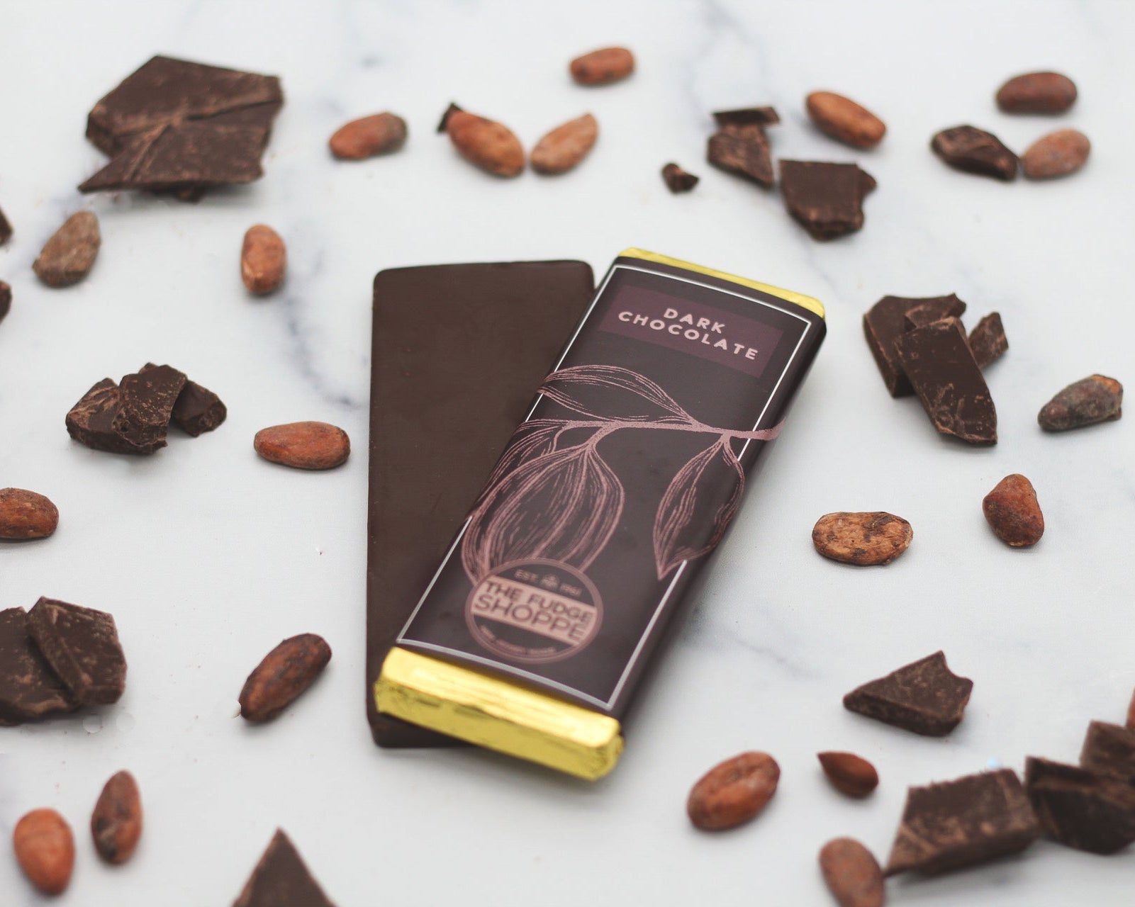 A dark chocolate bar with a gold wrapper, surrounded by scattered cocoa beans and broken pieces of chocolate on a marble surface.