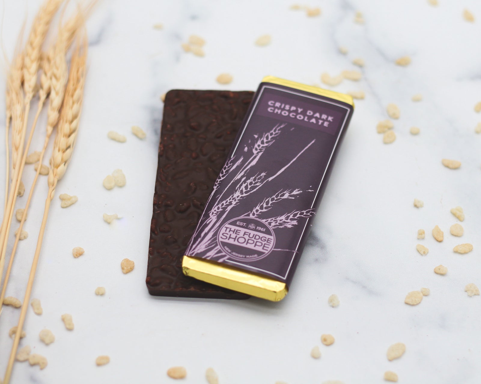 A dark chocolate bar mixed with rice crispies, presented on a marble surface with rice grains scattered around and a wheat sheaf in the corner.