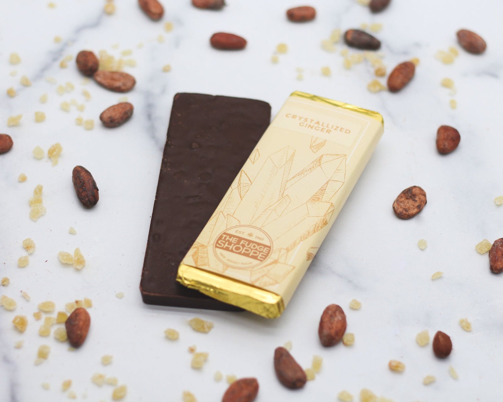 A dark chocolate bar with visible crystalized ginger cubes, presented on a marble surface with cocoa beans scattered around.
