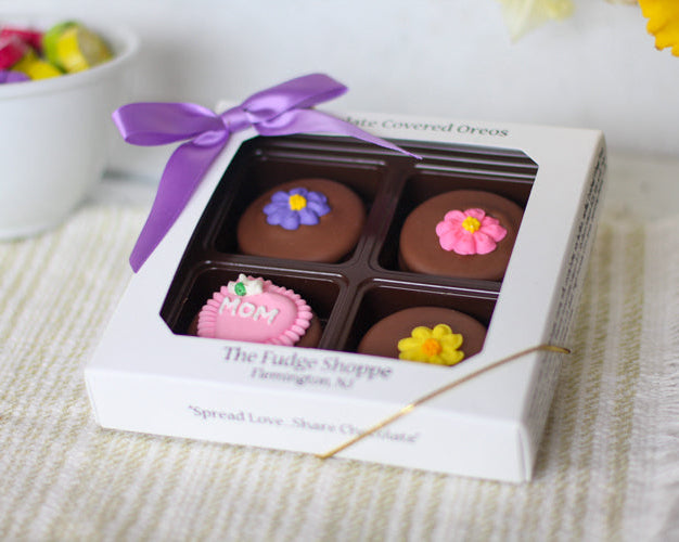 A box of Mother's Day Oreo cookies with four chocolate covered oreos decorated with colorful flower sugar pieces, presented in a white box with a purple ribbon.
