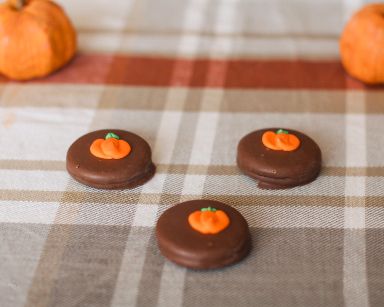 Three pumpkin-shaped chocolate treats decorated with orange frosting and a small green stem, arranged on a beige and brown plaid tablecloth with small orange pumpkins in the background.