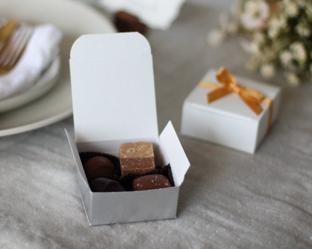 Small white box with chocolate candies on a gray surface with a plate and flowers in the background