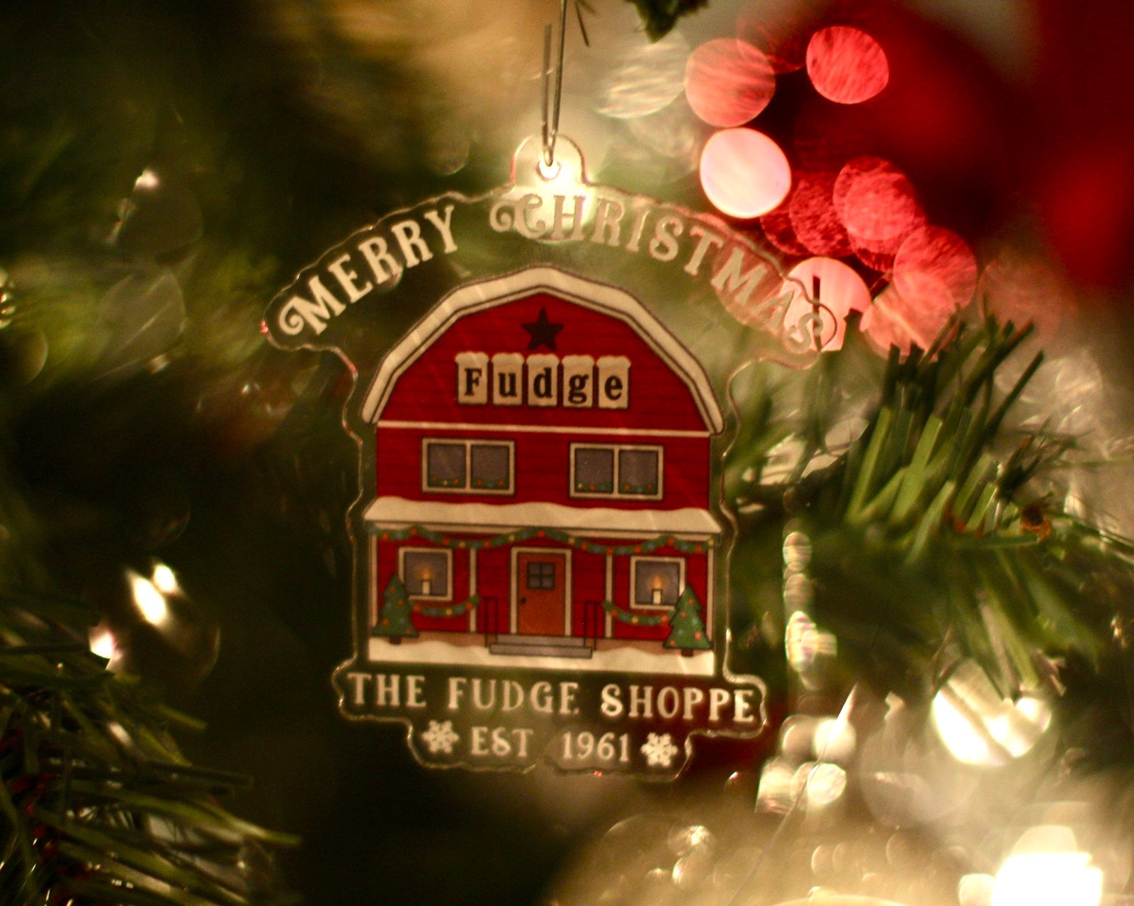 Christmas ornament of a red barn-style building with 'Merry Christmas' and 'The Fudge Shoppe' text, hanging on a tree.