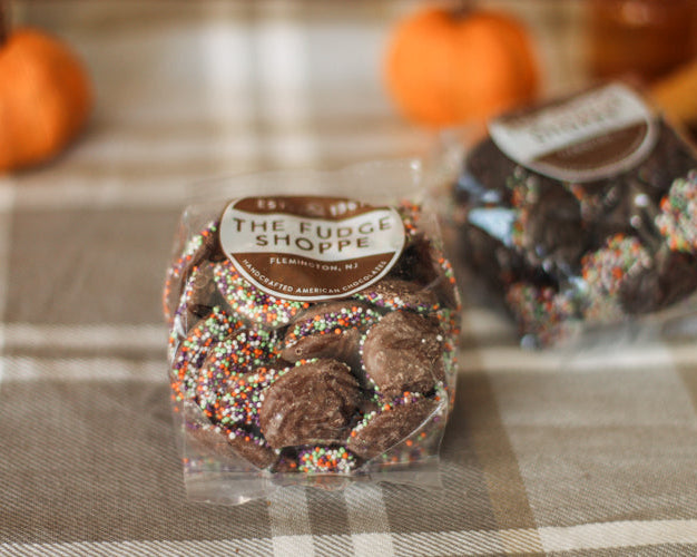 Chocolate non pareils with sprinkles in a clear package on a plaid tablecloth with pumpkins in the background.