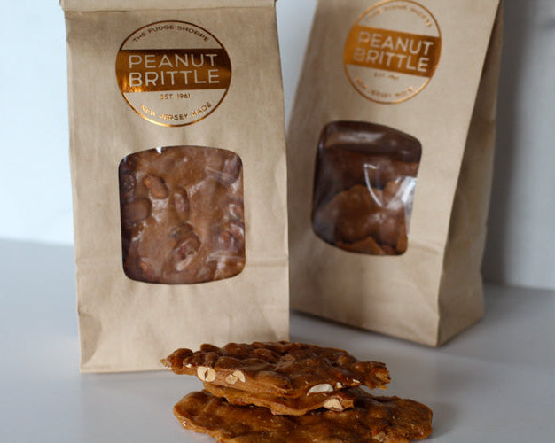 Packaged peanut brittle in brown paper bags with a clear window showing the product inside. Additional pieces of peanut brittle are displayed in front of the bags.