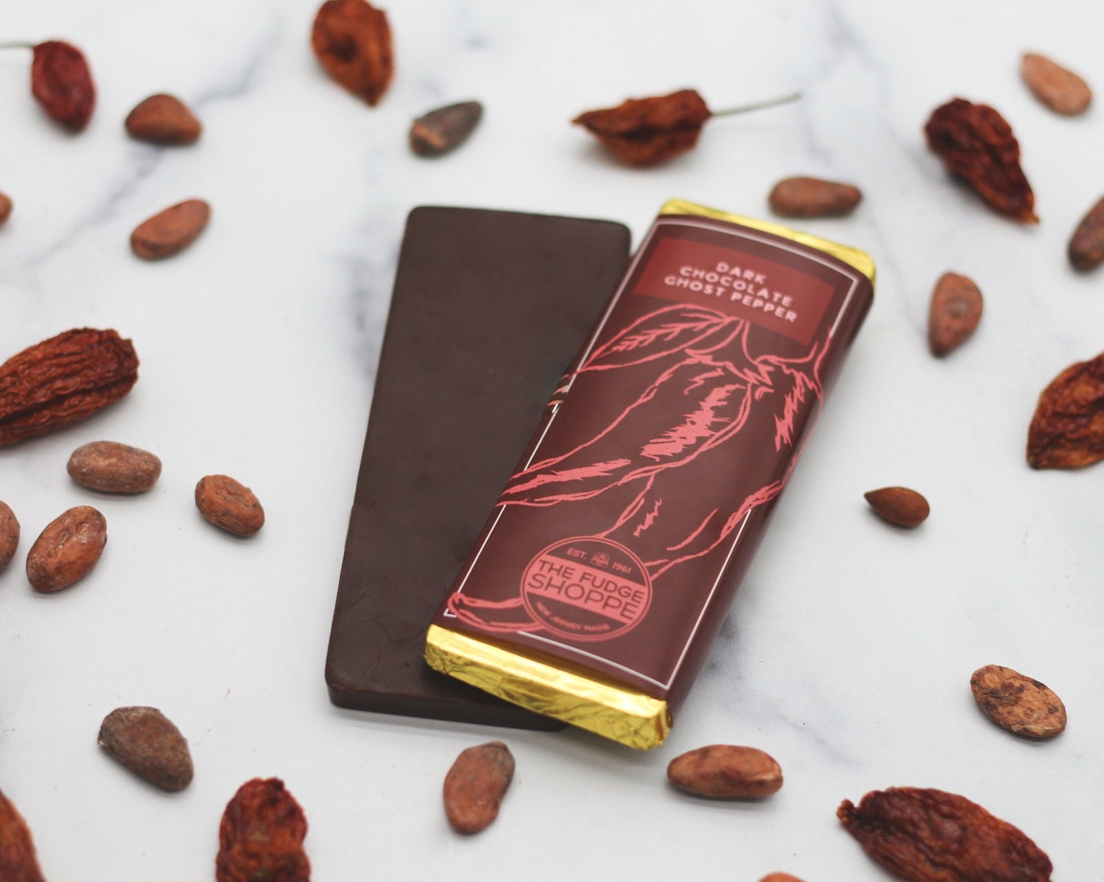 A dark chocolate bar with visible spicy ghost pepper particles, presented on a marble surface with scattered ingredients around it.