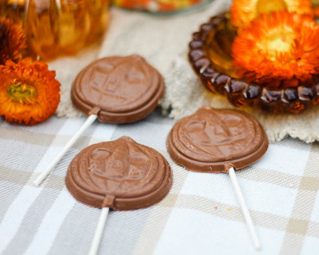 Chocolate lollipops with a jack o' lantern design, displayed on pops on a cloth surface with autumn-themed decorations around.