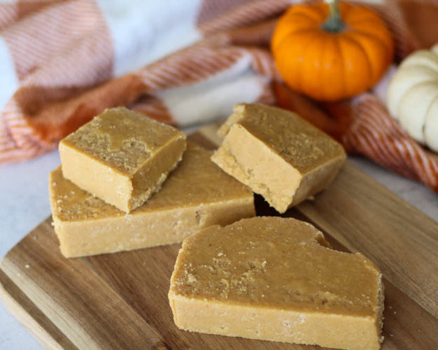 Three square pieces of pumpkin fudge on a wooden cutting board with a fall-themed textured background, including a pumpkin and autumn leaves.