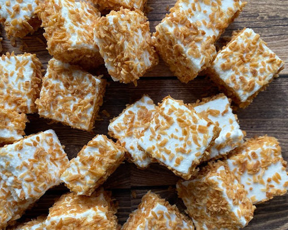 A group of toasted coconut marshmallows cut into squares and coated with toasted coconut flakes, displayed on a wooden surface.