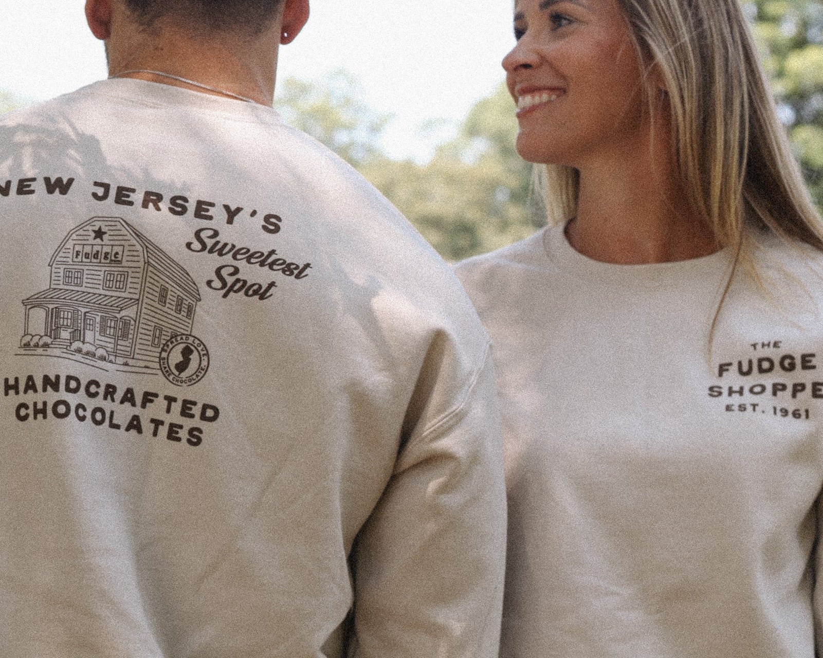 Two people wearing beige sweatshirts with text and a logo, standing outdoors.