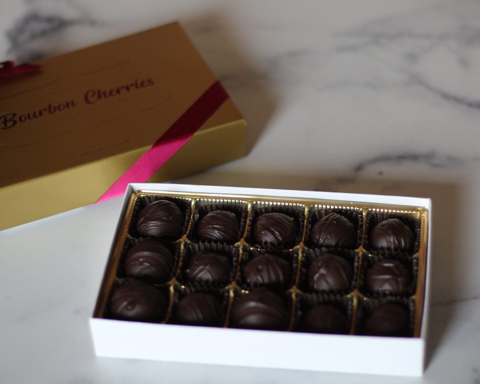 A box of bourbon cordial cherries with cherries coated in chocolate, arranged neatly inside a white box with a gold lid. The box is placed on a marble surface.