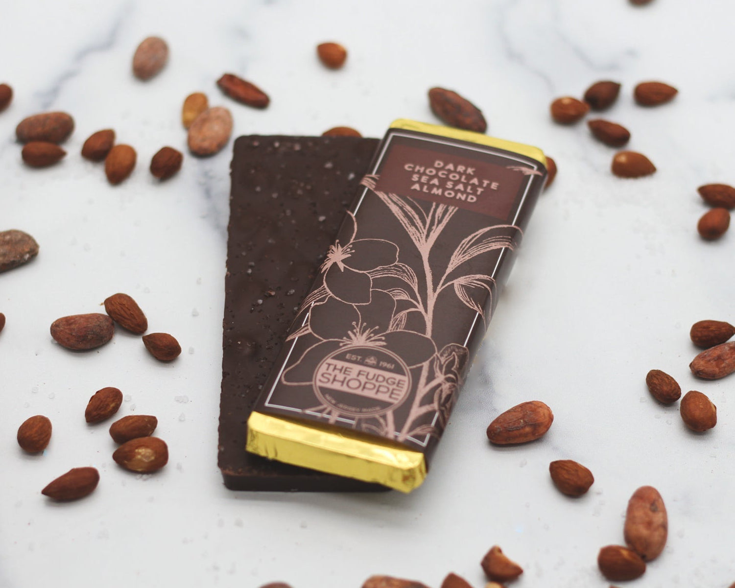 A dark chocolate bar with sea salt and almond pieces, presented on a marble surface with scattered almonds and cocoa beans.