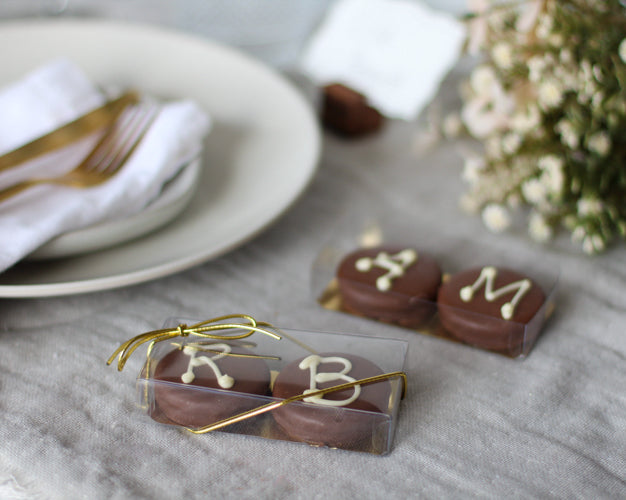 Chocolate candies with letters 'R', 'B', and 'M' on a gray surface with a plate and flowers in the background.