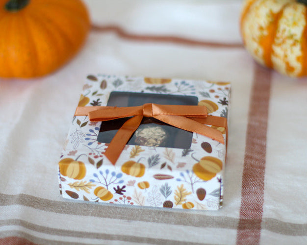 A small, decorated chocolate assortment box with a fall theme, featuring autumn colors and designs, presented on a cloth with pumpkins in the background.