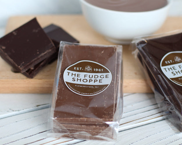 Packaged homestyle chocolate from The Fudge Shoppe on a wooden board with a bowl of fudge in the background.