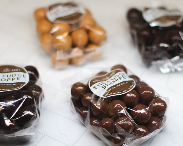 Packages of chocolate malt balls from The Fudge Shoppe on a white surface.