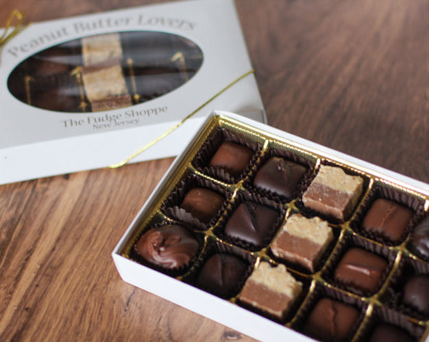 A box of assorted chocolate peanut butter treats, including pieces in square and round shapes, displayed in an open box with a lid that reads 'Peanut Butter Lovers' on a wooden surface.