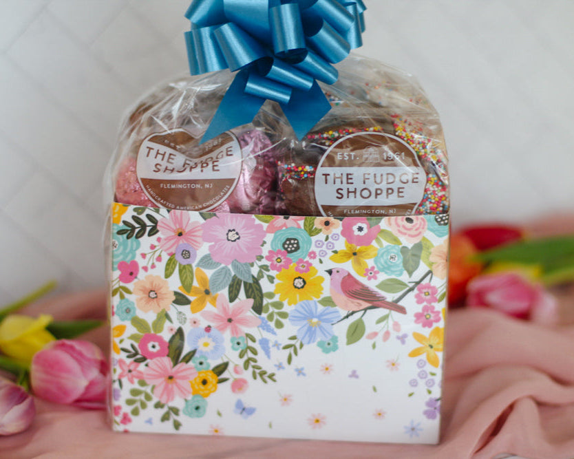 Packaged chocolates from The Fudge Shoppe with a floral design and blue bow on a white background.