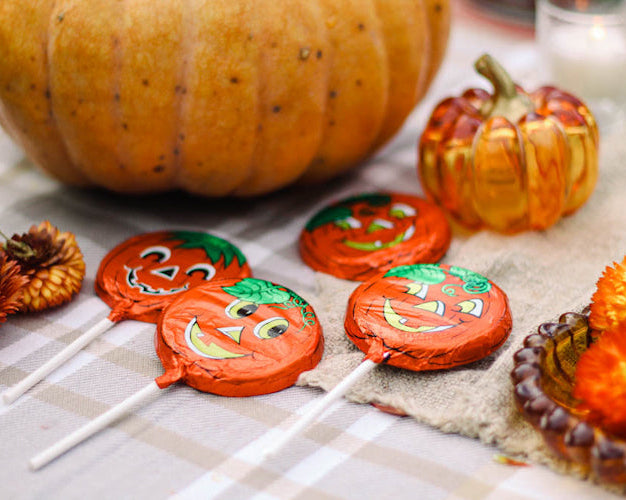 Milk chocolate pops with foil wrappers, designed to look like pumpkins with Halloween-themed patterns, displayed on a table with autumn-colored decorations around.