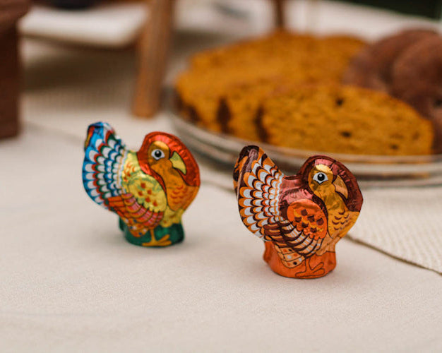 Small foiled chocolate turkeys with colorful details, placed on a table with various baked goods in the background.