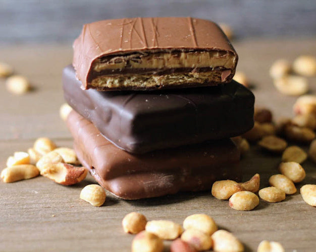 Stack of chocolate covered peanut butter grahams with visible layers of chocolate and peanut butter, surrounded by peanuts on a wooden surface.