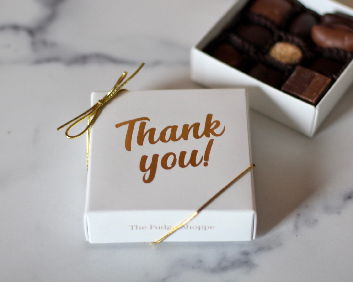 A white box with 'Thank you' printed in gold, containing an assortment of chocolate candies, tied with a gold ribbon.